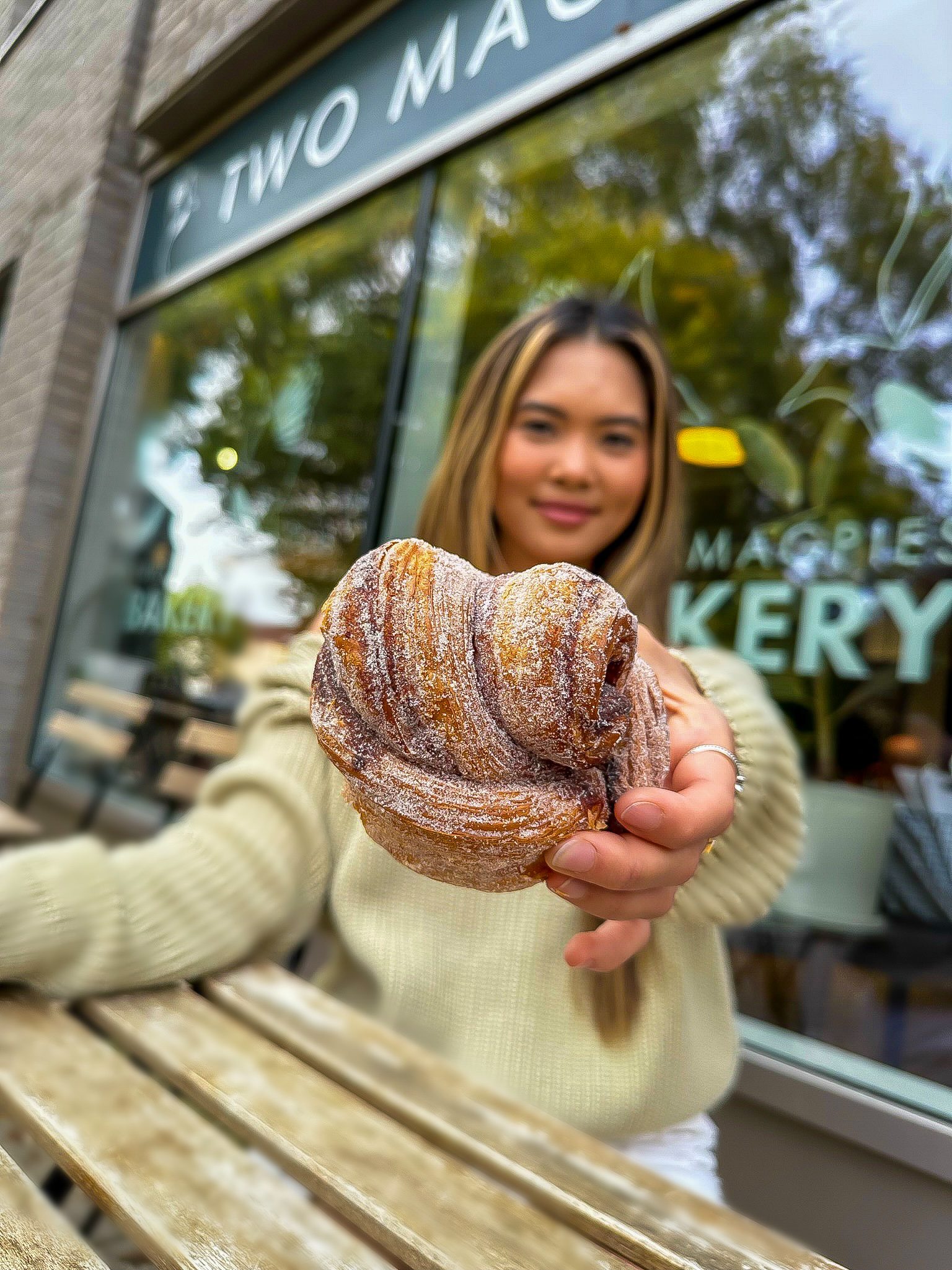 a young woman with dark hair and skin holding a cinnamon swirl in her hand outside of a bakery.